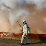 A worker in a full protective suit fumigates an area outdoors, spraying pesticide.